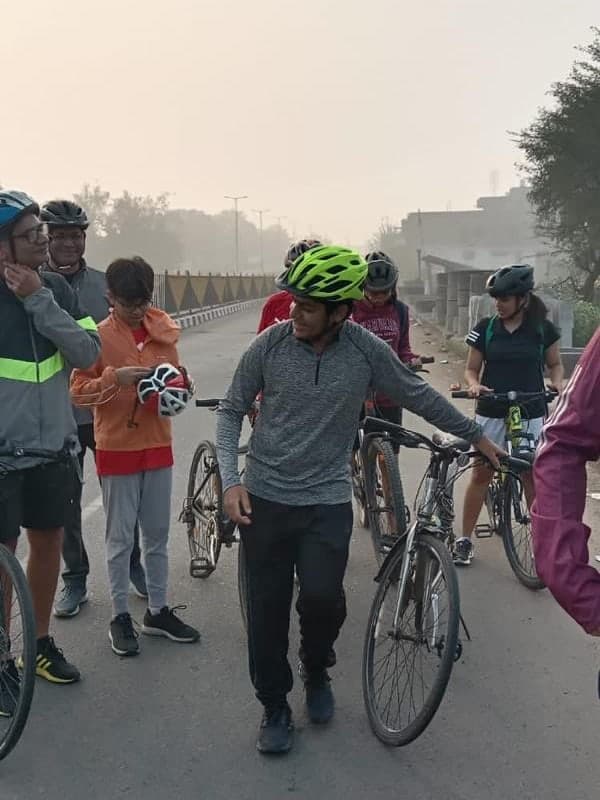 Tarush in a green helmet walking his bike on a road, with other cyclists around him on a hazy day.