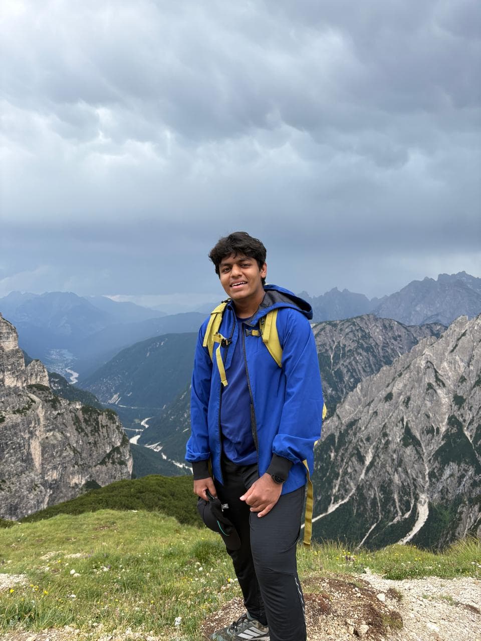 Tarush on a grassy ridge in a blue rain jacket and yellow backpack, with jagged mountains and a valley behind him under heavy cloud.