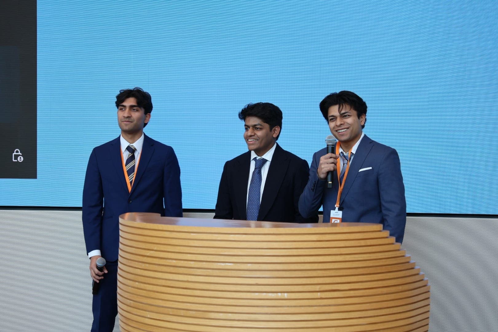 Tarush on stage in a dark suit between two others, one holding a microphone, in front of a large blue screen.