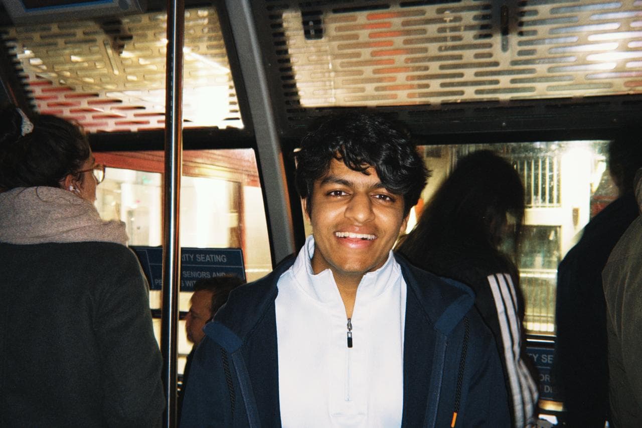 Tarush grinning on a crowded transit car, in a navy jacket over a white half-zip.